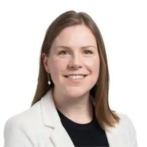 A woman with straight light brown hair, wearing a white blazer, black top, and a single dangling earring, smiles at the camera against a plain white background.