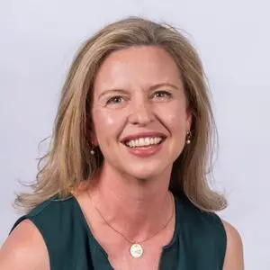 A woman with shoulder-length blonde hair, wearing a sleeveless dark green top, small earrings, and a necklace, smiling at the camera against a plain light background.