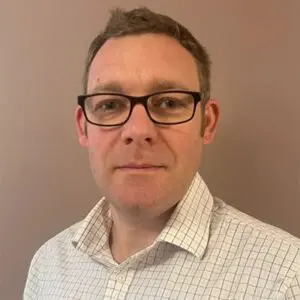 A man with short hair and glasses wearing a light checked shirt stands against a plain, beige background.