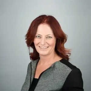 Woman with shoulder-length red hair, wearing a grey and black jacket, smiles at the camera against a plain light grey background.