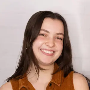A young woman with straight brown hair, wearing a brown sleeveless top, smiles in front of a white brick wall.