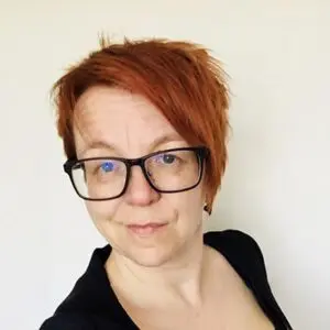 Person with short red hair and black glasses looks at the camera, standing in front of a plain white background, wearing a black top.