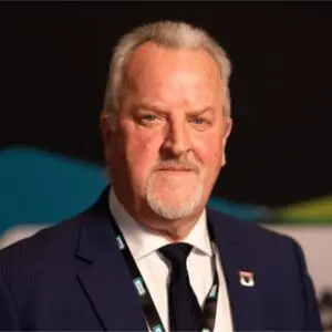 Middle-aged man with grey hair and beard wearing a navy suit, white shirt, and dark tie, standing indoors with a lanyard round his neck.