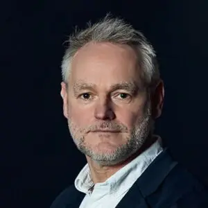 A middle-aged man with grey hair and beard wearing a white shirt and dark jacket poses against a dark background.