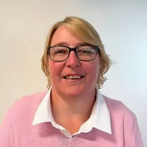 A person with short blonde hair and glasses, wearing a white collared shirt under a pink jumper, is facing the camera with a neutral expression against a plain light background.