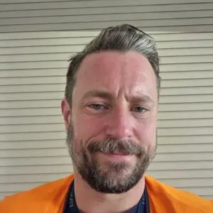 A man with short brown hair and a beard is wearing an orange shirt and smiling in front of beige horizontal blinds.