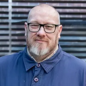 A man with glasses, a short beard, and a blue buttoned jacket stands outdoors in front of slatted metal panels.