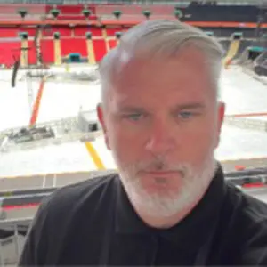 A man with short white hair and beard, possibly Mike Chalmers, wearing a black shirt stands in a stadium with red seats and a partially covered pitch in the background, capturing the atmosphere as anticipation builds for events leading up to 2026.