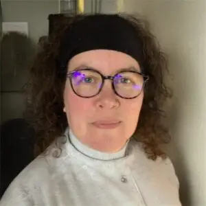 A person with curly hair, glasses, and a black hairband looks towards the camera, wearing a light-coloured polo-neck jumper, standing indoors.