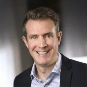 Smiling man in a checked shirt and dark blazer, with short brown hair, posing indoors against a blurred background.