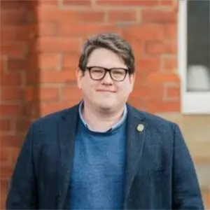 A person with short brown hair and glasses is standing outdoors in front of a brick wall, wearing a blue jumper and a dark blazer.