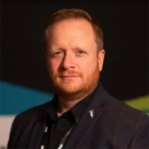 A man with short red hair and a beard wearing a dark checked blazer and black shirt stands in front of a dark background.