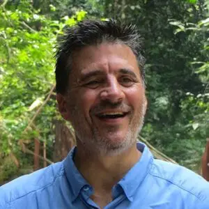 A man with short dark hair and a beard smiles whilst standing outdoors surrounded by green trees, wearing a light blue collared shirt.