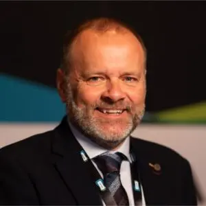 A middle-aged man with a beard in a suit and tie is smiling at the camera, wearing a lanyard and a lapel pin, with a dark background behind him.