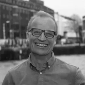 A man with glasses and a button-up shirt smiles outdoors in front of a blurred cityscape and water.