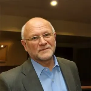 A middle-aged man with glasses, a grey beard, and a suit jacket over a blue shirt, standing indoors under warm lighting.