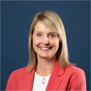 Woman with shoulder-length blonde hair wearing a coral blazer and white top, smiling, posed in front of a plain dark blue background.