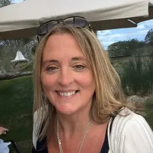 Smiling woman with long blonde hair wearing sunglasses on her head, a white jumper, and a silver necklace, outdoors with greenery and a white marquee in the background.