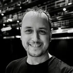Smiling man in a black shirt stands indoors; background features industrial ceiling and lighting, suggesting a theatre or studio setting. Black-and-white photo.