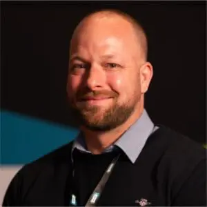 A man with a beard and shaved head, wearing a dark jumper over a collared shirt, smiles whilst facing the camera against a dark background.