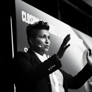 Person in a suit with a headset microphone gestures with both hands while speaking on stage in front of a large presentation screen, delivering an engaging talk about Satia Rai’s vision for 2026.