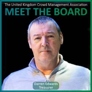 Portrait of a man in a light grey zip-up shirt with text identifying him as Darren Edwards, Treasurer and one of the Board Members of the United Kingdom Crowd Management Association board.