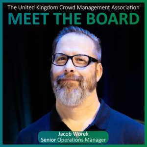 A man with glasses and a grey beard stands in front of a dark background. Text reads: Meet the Board Members. Jacob Worek, Senior Operations Manager. The United Kingdom Crowd Management Association.