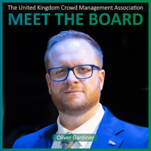 A man with glasses and a beard wears a blue suit and tie. Text above reads The United Kingdom Crowd Management Association Meet the Board Members. Name tag: Oliver Gardiner.