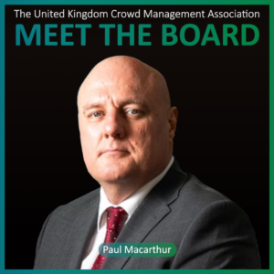 A man in a suit and tie poses against a black background. Text above reads The United Kingdom Crowd Management Association MEET THE BOARD. Name below: Paul Macarthur, highlighting one of the distinguished Board Members.