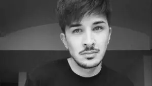 A young man with short dark hair, a trimmed beard, and moustache looks at the camera. He is wearing a dark shirt. The background is plain and in greyscale.