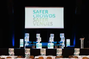 A conference stage with a large screen displaying SAFER CROWDS SAFER VENUES, two chairs, lectern, and event signage. Audience tables in the foreground.