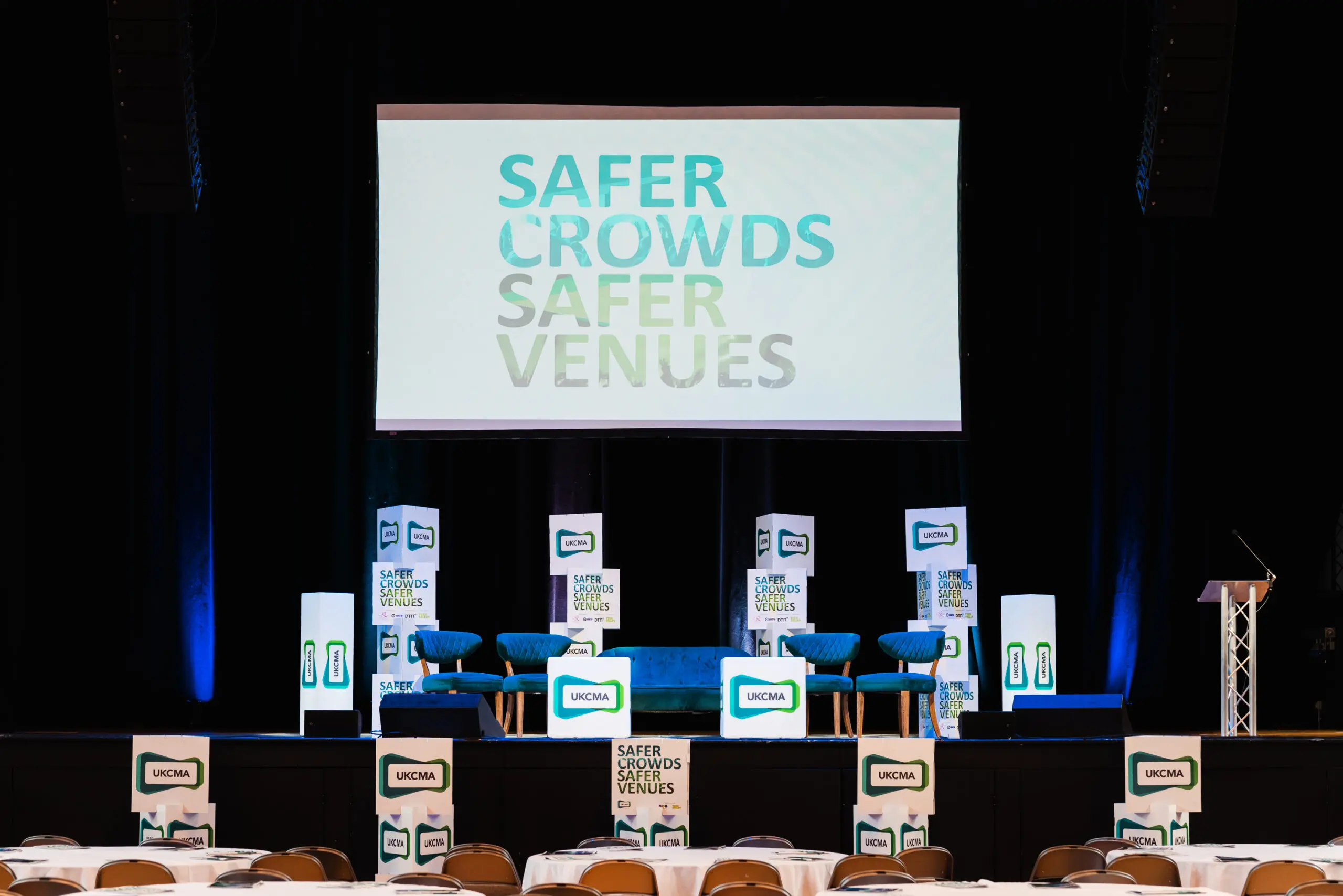 A conference stage with a large screen displaying SAFER CROWDS SAFER VENUES, two chairs, lectern, and event signage. Audience tables in the foreground.