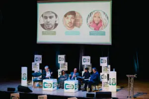 Five panellists sit on a stage with UKCMA signage, while three faces and names are displayed on a large screen above them.