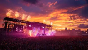 A large crowd watches a concert at sunset with fireworks and colourful stage lights illuminating the sky.