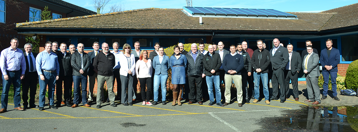 A group of about 25 adults stands in a queue outside a building with solar panels on the roof, under a clear blue sky.